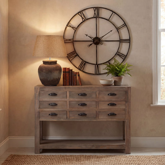 Wooden console table with lamp, books, and decorative items against a wall with a large clock.