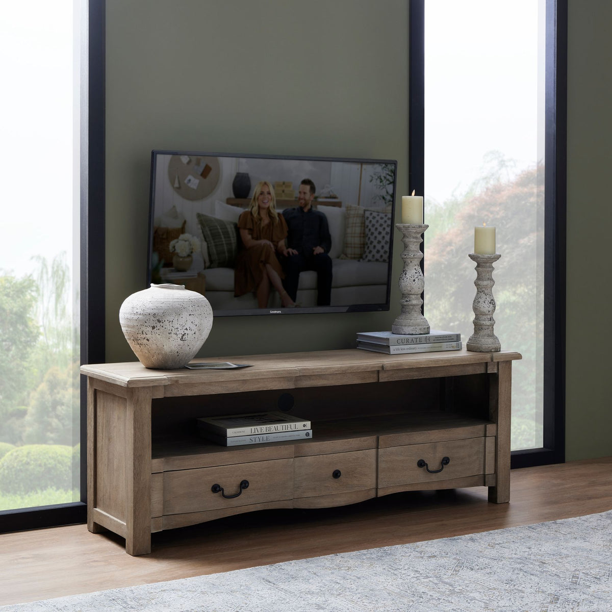 Wooden TV stand with a television displaying two people, surrounded by decorative items in a room with large windows.