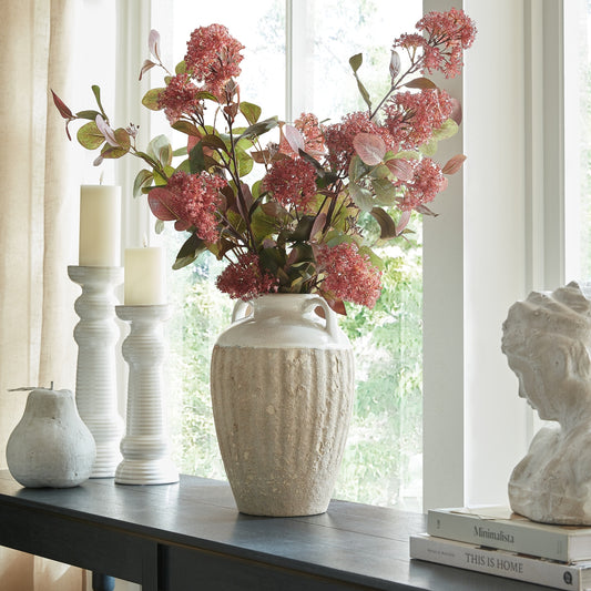 Decorative setup with a textured vase holding pink flowers, candles, and books on a table.