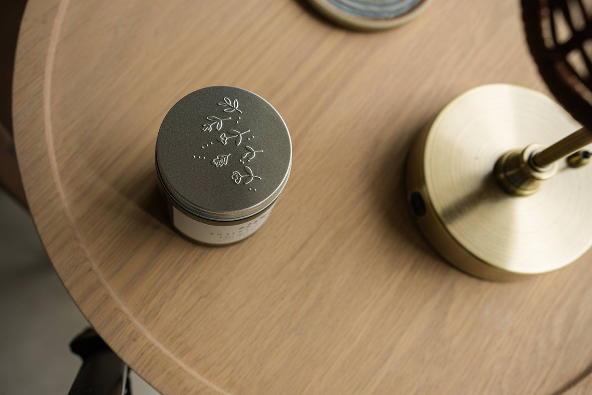 Embossed silver Embers & Roots candle lid beside a candle on a wooden surface