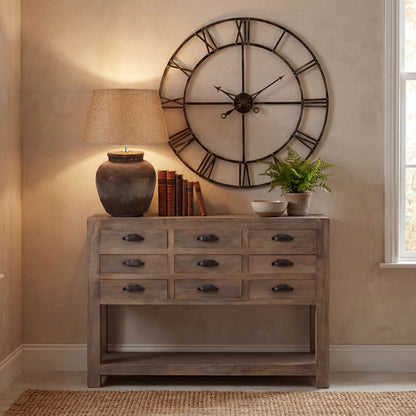 Wooden console table with lamp, books, and decorative items against a wall with a large clock.