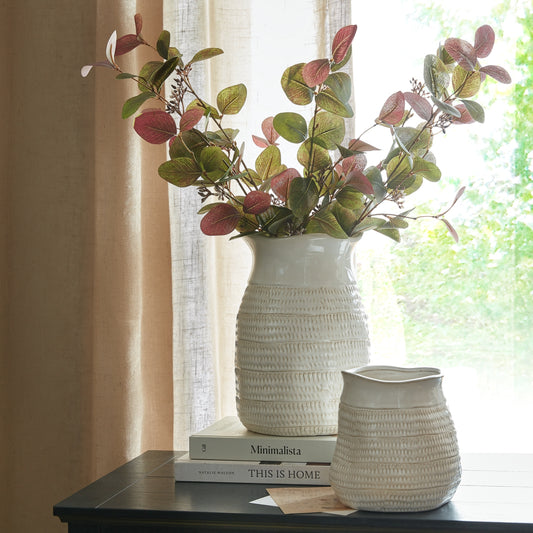 White textured vases with a plant on a table next to books, with a window and curtain in the background.