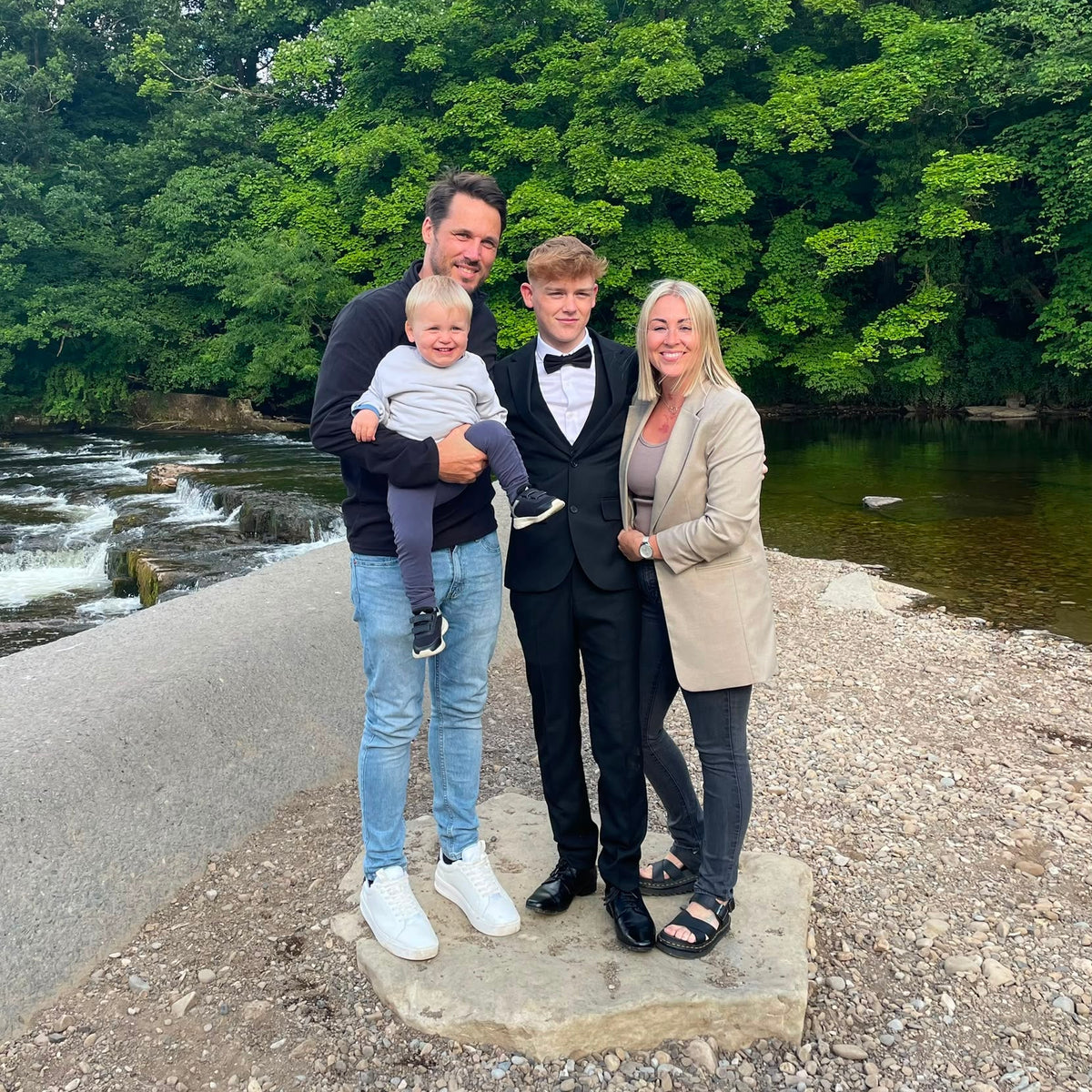 A family of four posing outdoors on a stone slab by a river, with a man holding a baby, a boy in a black tuxedo, and a woman in a beige blazer, set against a backdrop of lush green trees and flowing water.