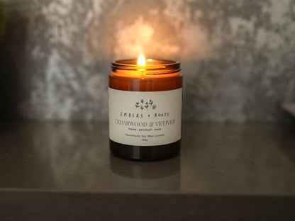 Lit Embers & Roots candle in an amber glass jar against a textured wall