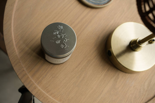 Embossed silver Embers & Roots candle lid beside a candle on a wooden surface